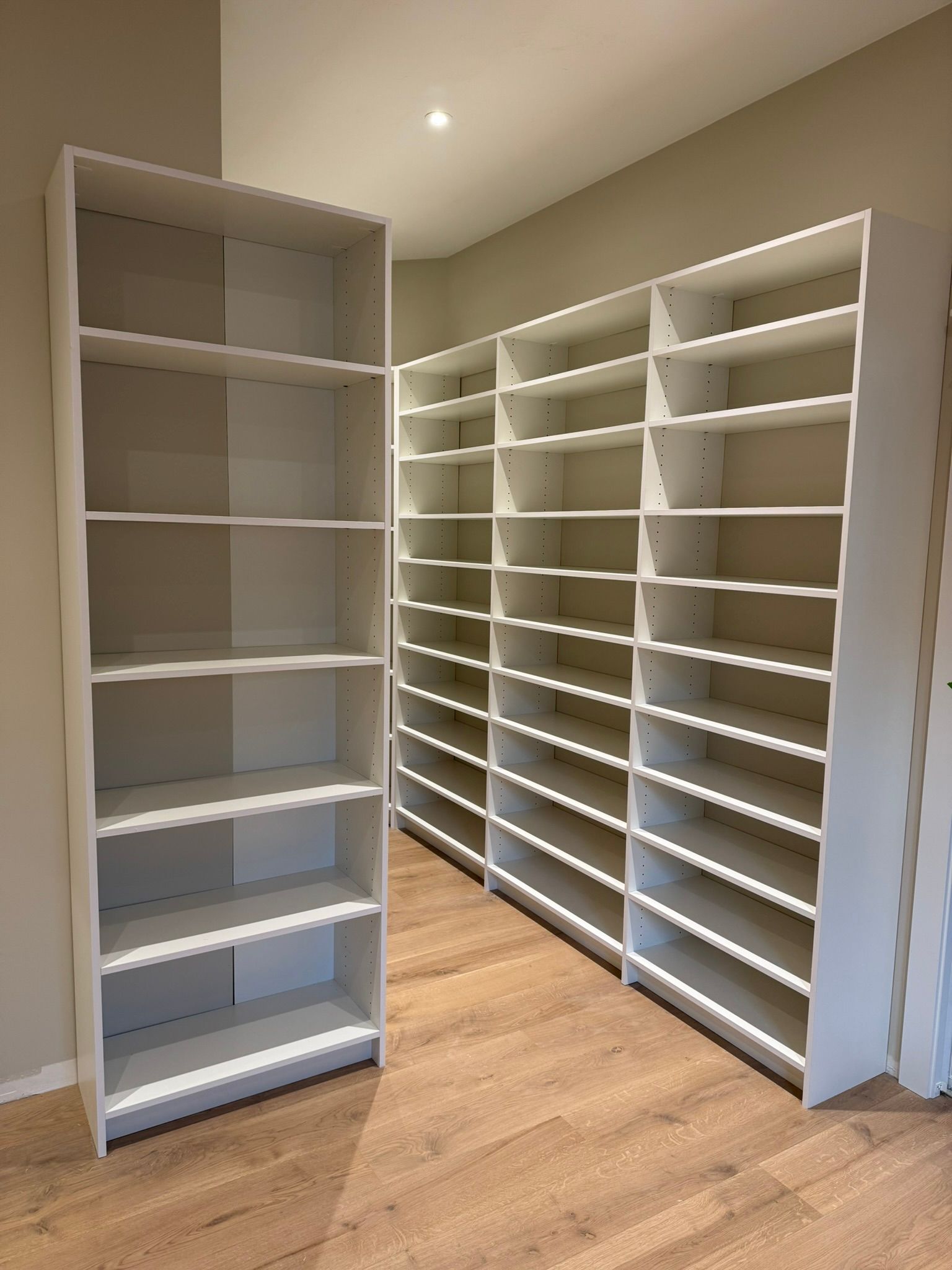 Three white bookshelves standing against a beige wall and hardwood floor.