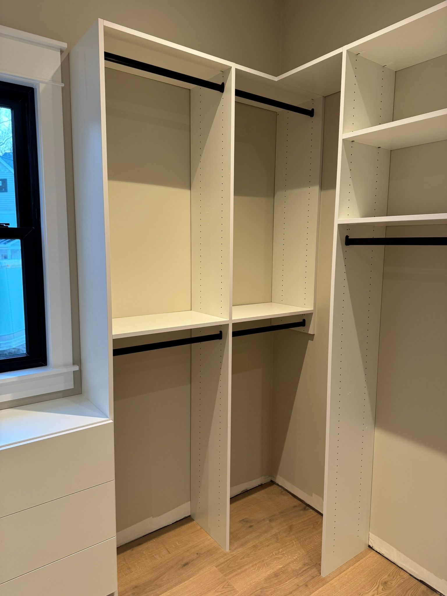White built-in closet with shelves and hanging rods. Light wooden floor, corner design, near a window.