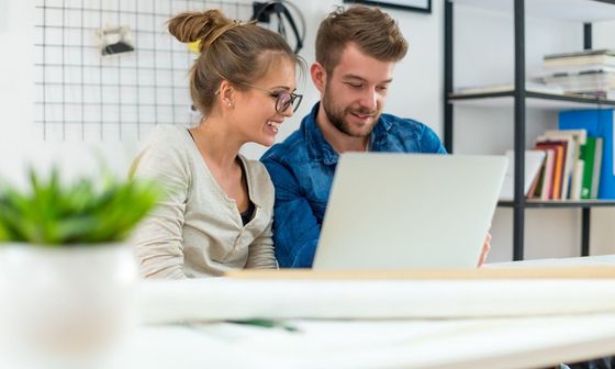 Two colleagues collaborate on a laptop in a bright, modern office space with a bookshelf in the background.