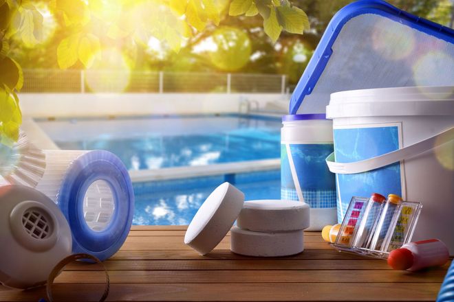 Pool cleaning supplies: filter, chlorine tablets, net, chemicals, and water test kit on wooden deck by a pool.