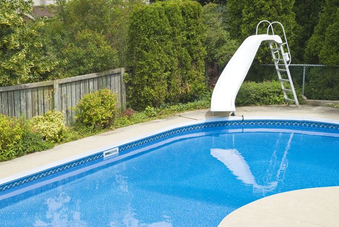 Swimming pool with a white slide and ladder, surrounded by greenery and a fence.