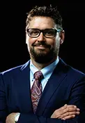 Smiling bearded man in glasses wearing a navy suit and patterned tie, arms crossed against a dark background