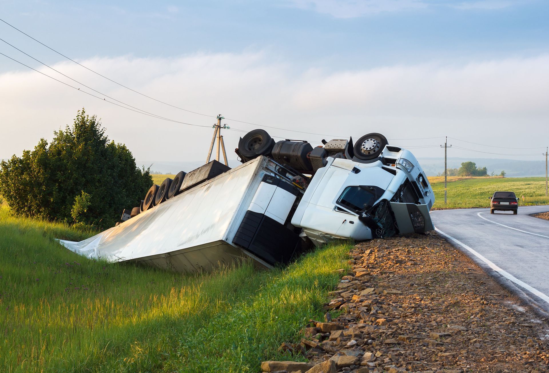 Overturned white truck on a rural roadside beside a wet highway at dawn