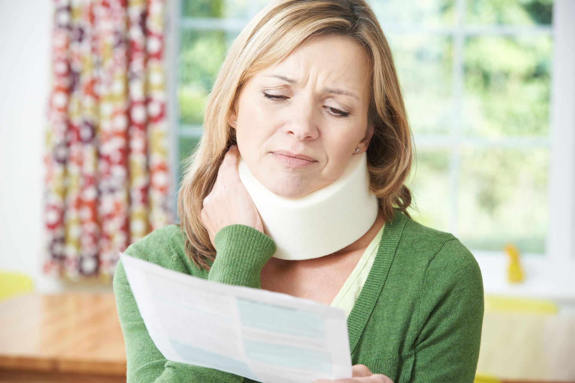 Woman in green sweater wearing a neck brace, reading a document at home with a pained expression
