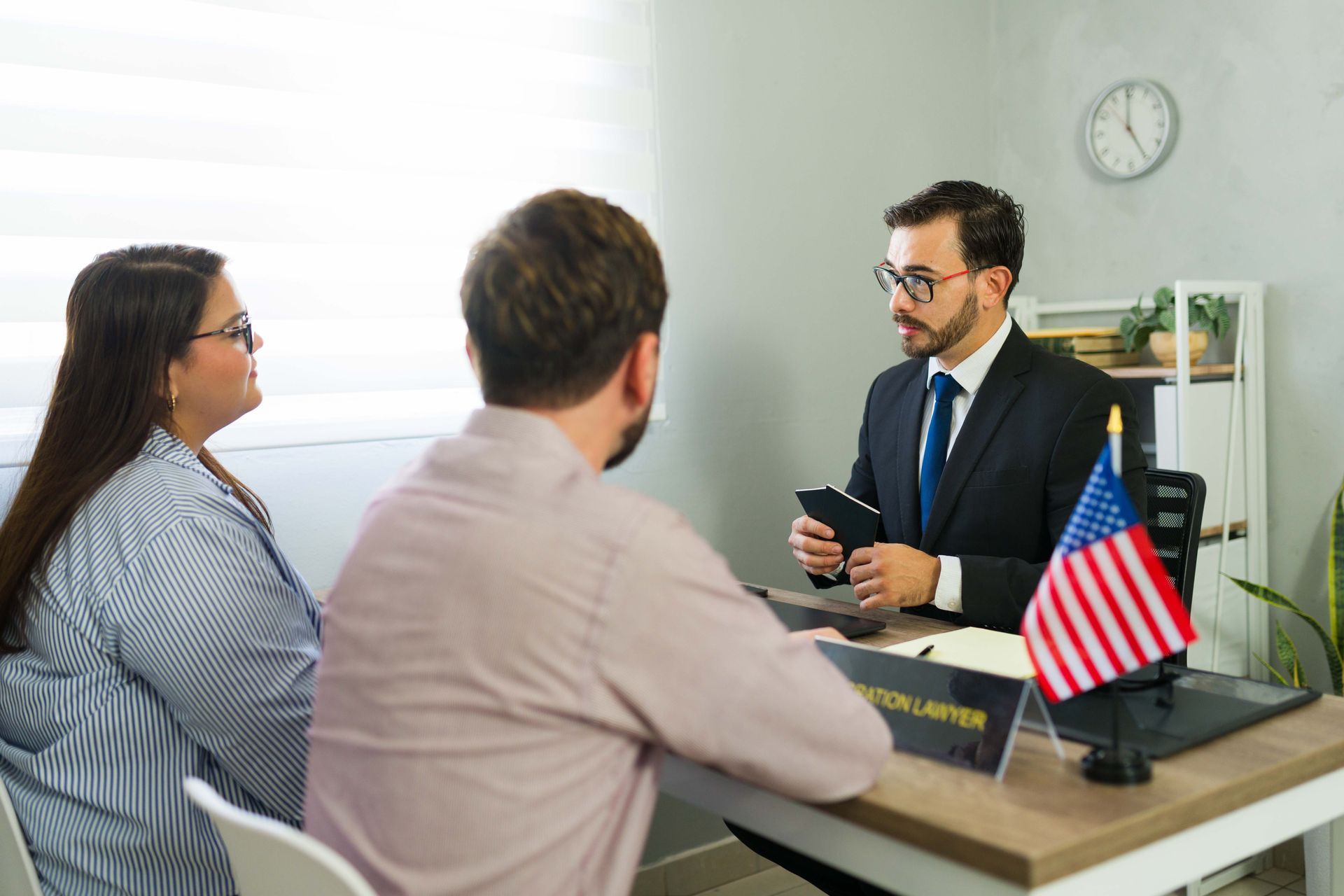 Two people meeting with a suited man at a desk, with a small American flag and clock in the office.