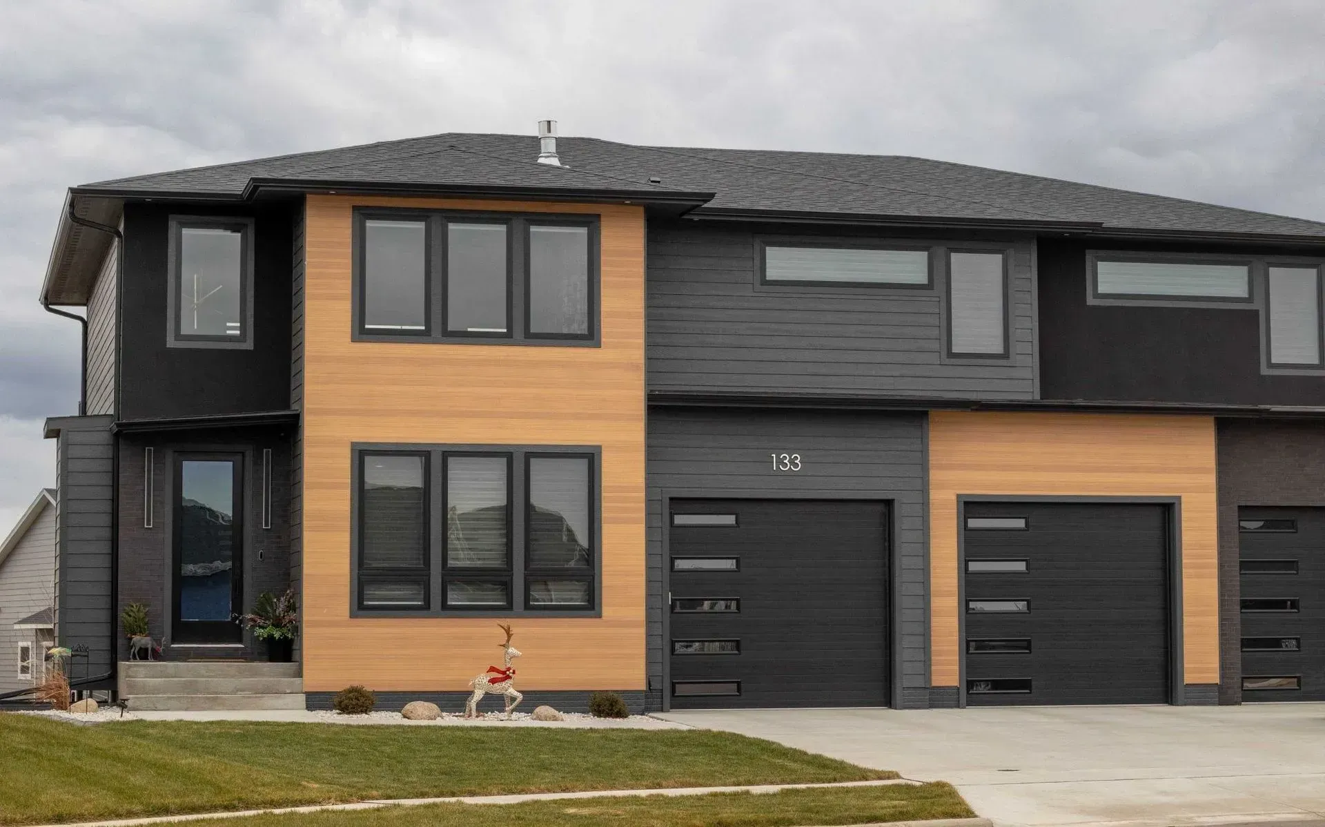 Modern two-story townhomes with black and tan siding. Two have attached garages, a green lawn, and a cloudy sky.