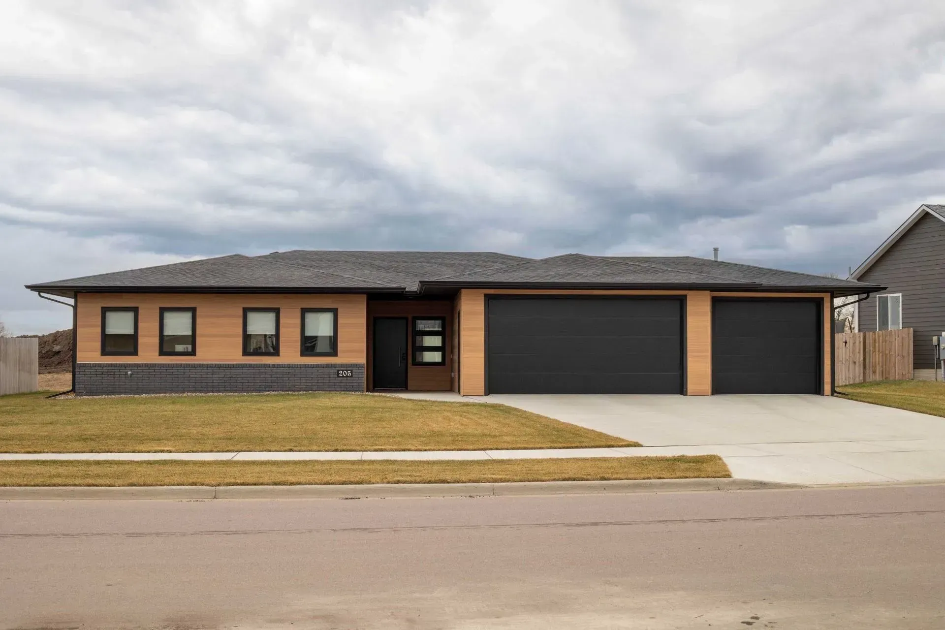 Modern one-story house with brown and black exterior, three-car garage, and cloudy sky.