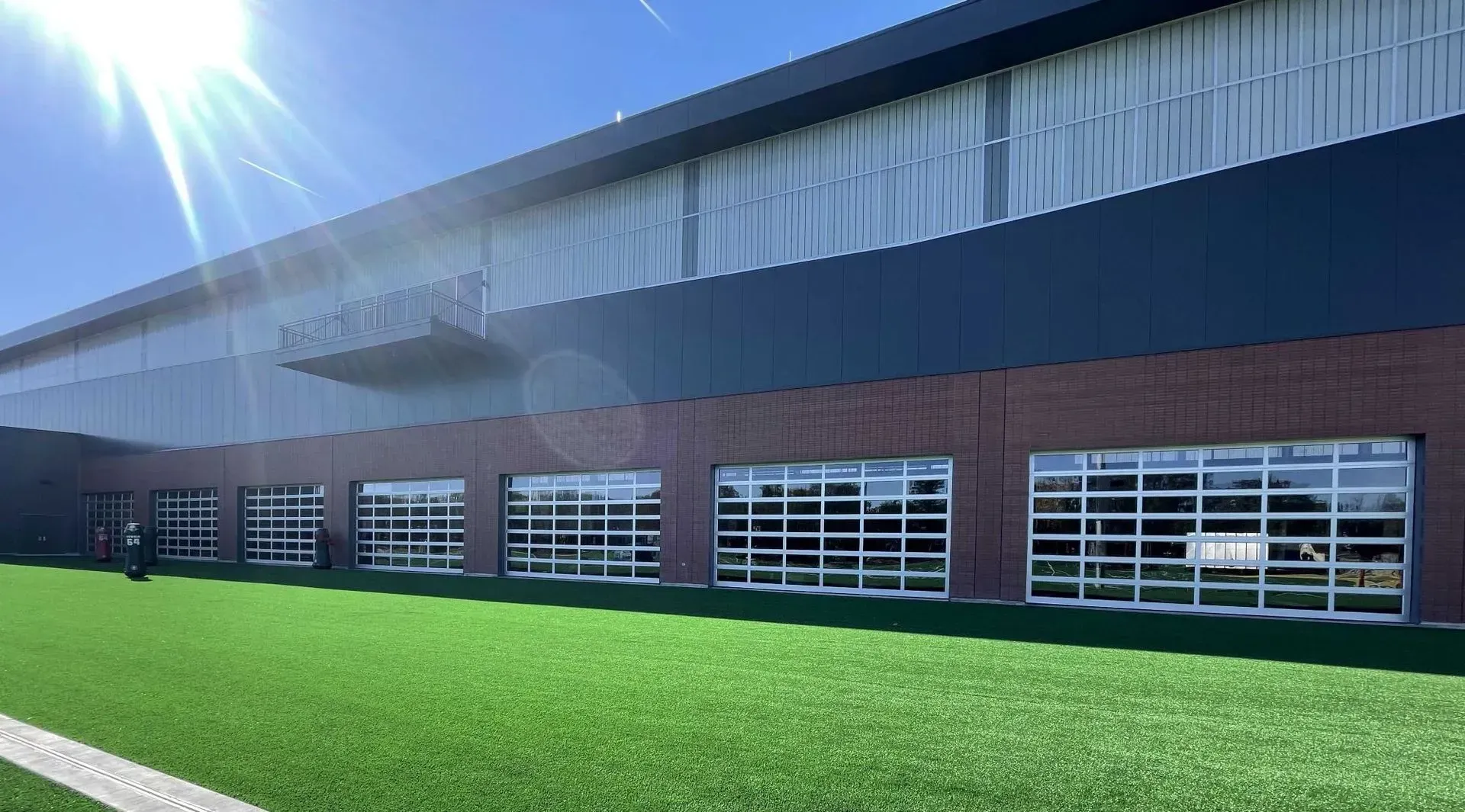 Exterior of a modern building with glass garage doors, brick and black panels. Artificial turf lawn. Sunny day.