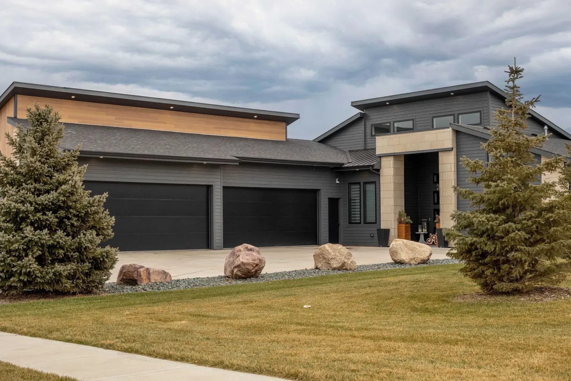 Modern grey house with attached garage, two-tone roof, and front lawn with decorative rocks and evergreens.