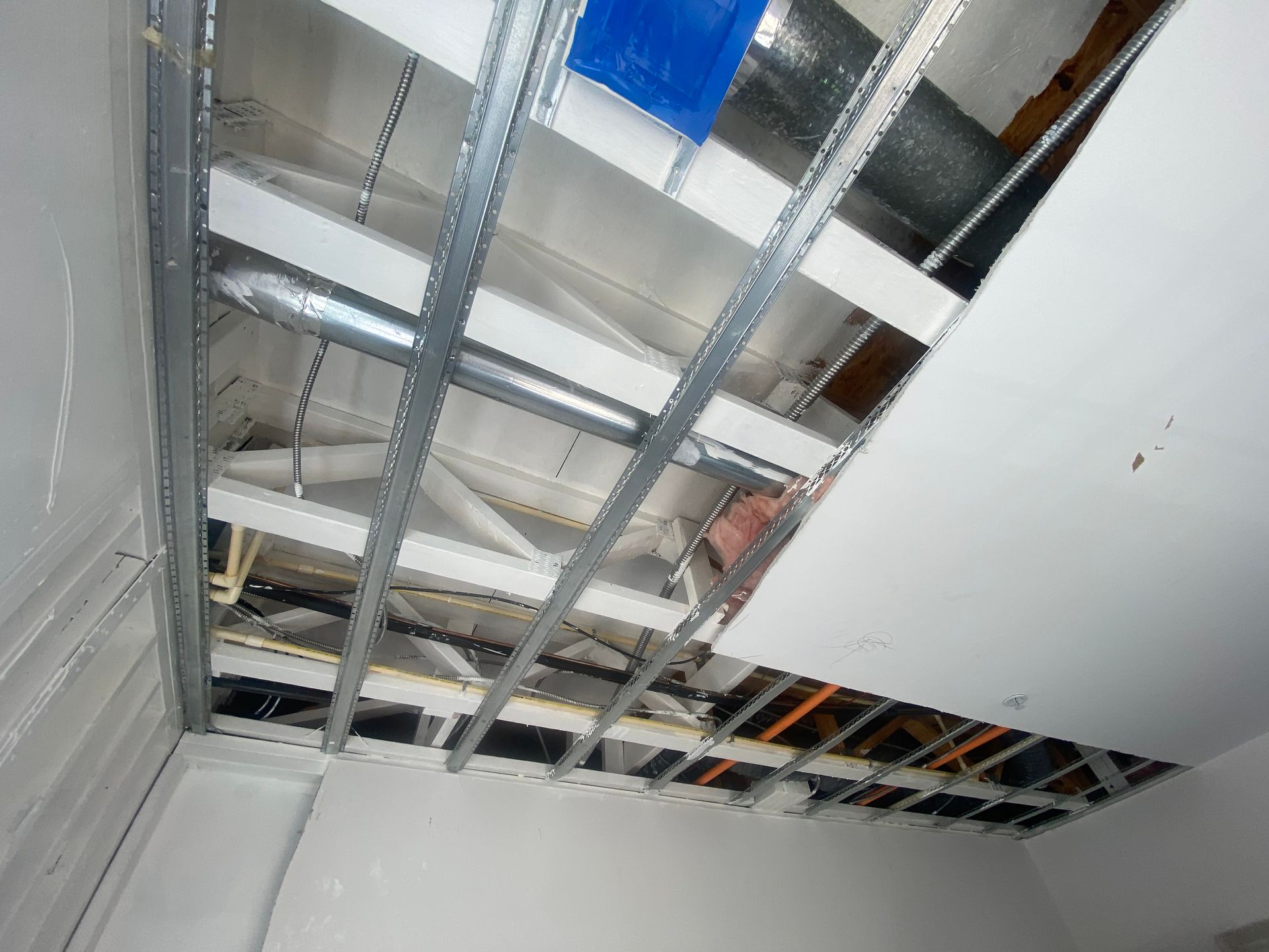 An unfinished ceiling with metal framing, exposed ductwork, pipes, and partial drywall installation.