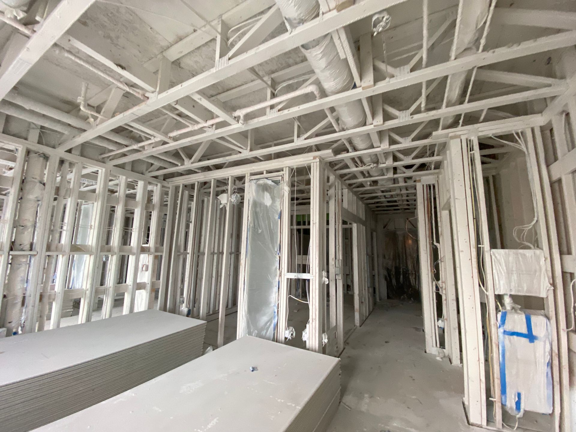 Interior of an unfinished building under construction with exposed metal framing, ceiling ducts, and drywall panels.