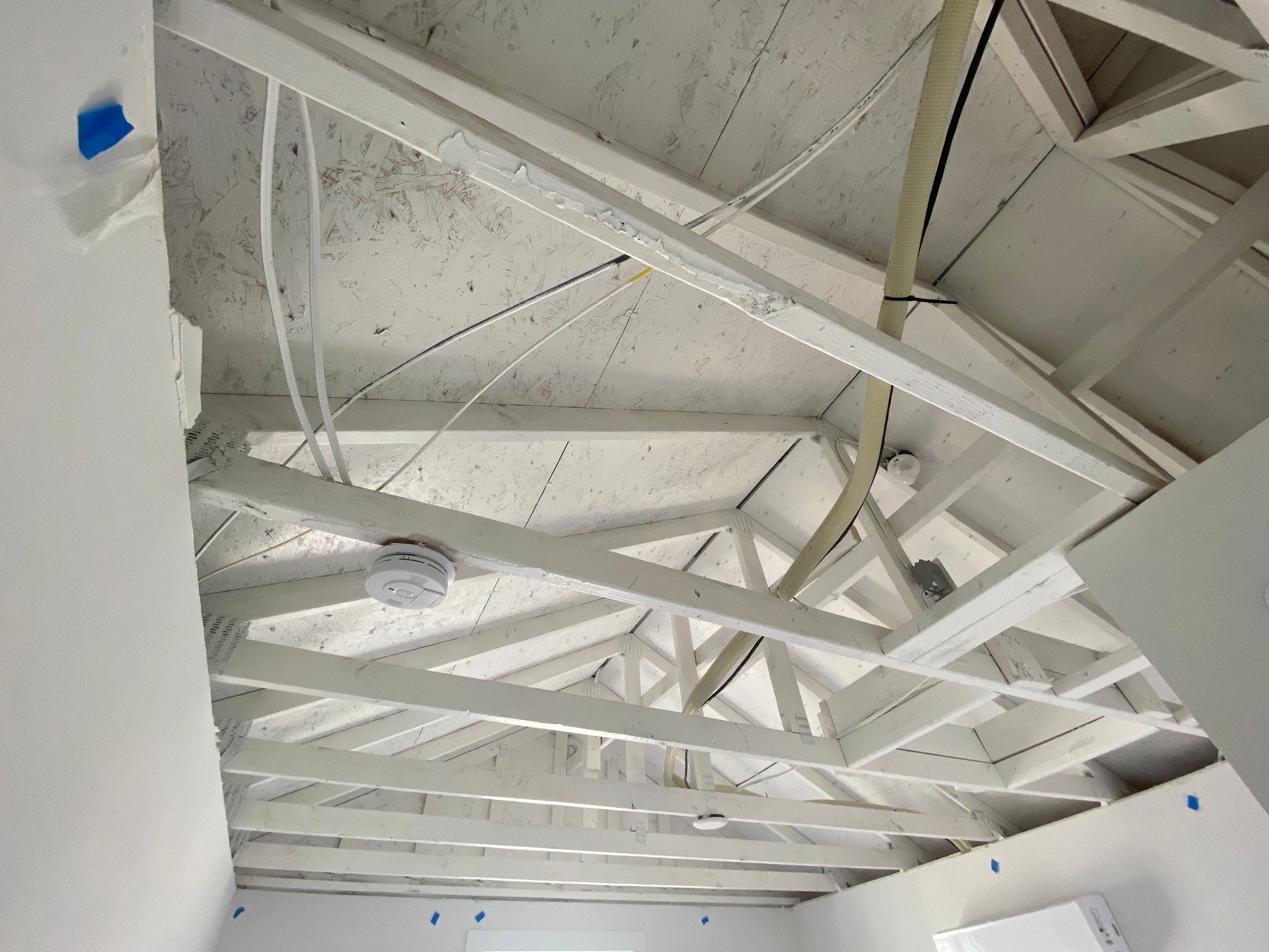 Low-angle view of a white, exposed timber truss roof frame above an unfinished interior room with ceiling lights.