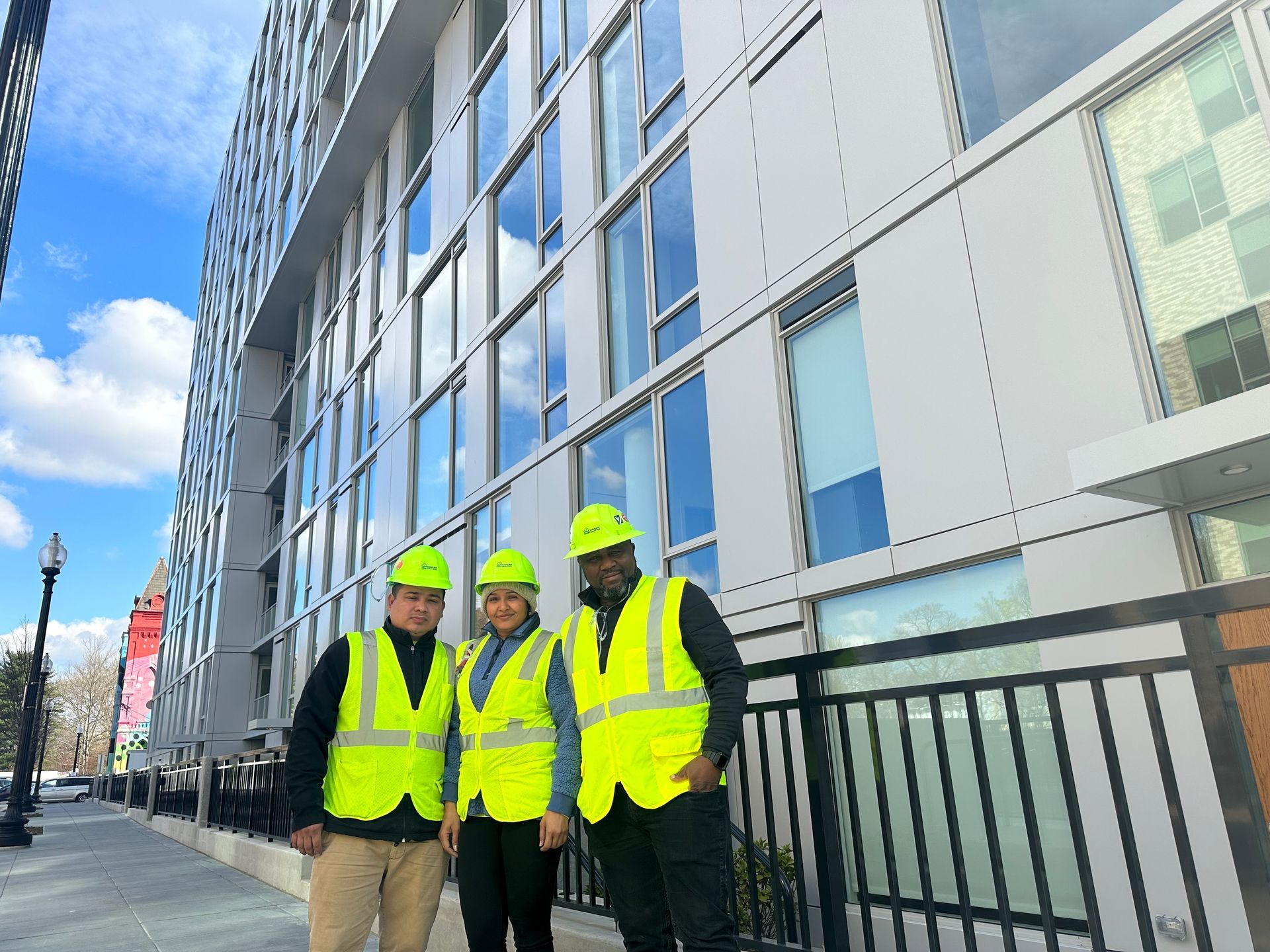 Three people in hard hats and high-visibility vests stand smiling in front of a modern glass and metal building.