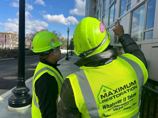 Two construction workers in neon yellow vests and hard hats inspect the exterior of a building, one pointing at windows.