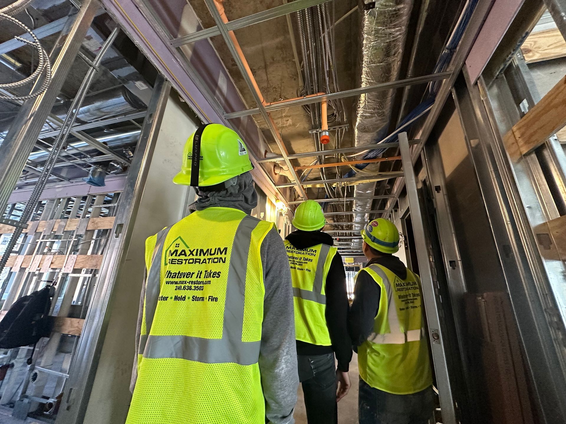 Three workers in safety gear walk through a construction corridor with exposed framework and ceiling ductwork.
