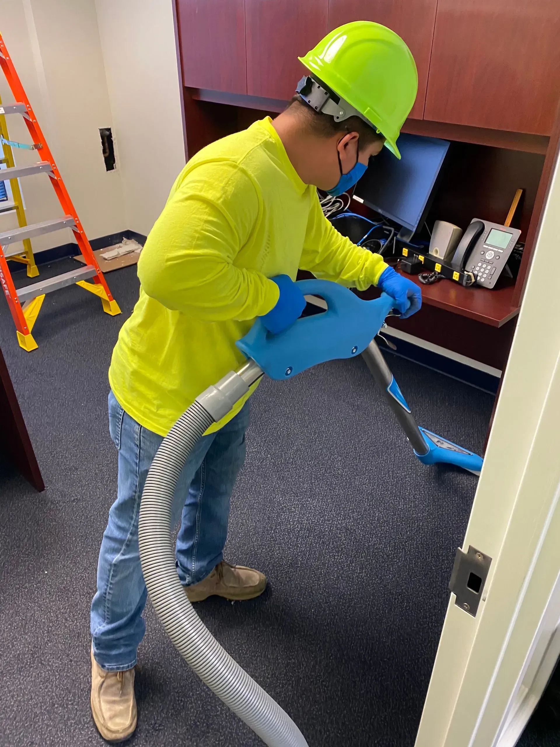Worker in a yellow shirt and neon hard hat vacuums a dark-carpeted office room.