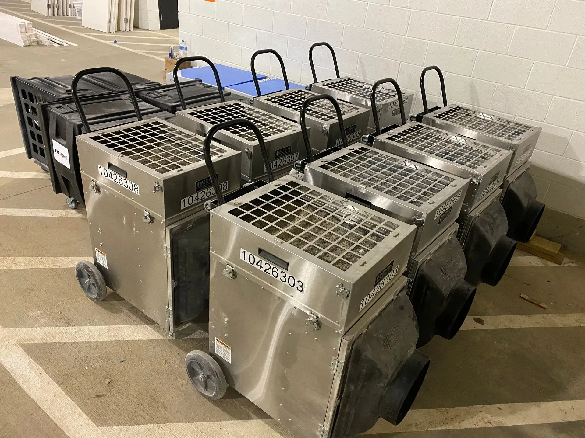 A row of metallic industrial dehumidifiers parked in a storage area on a paved floor.