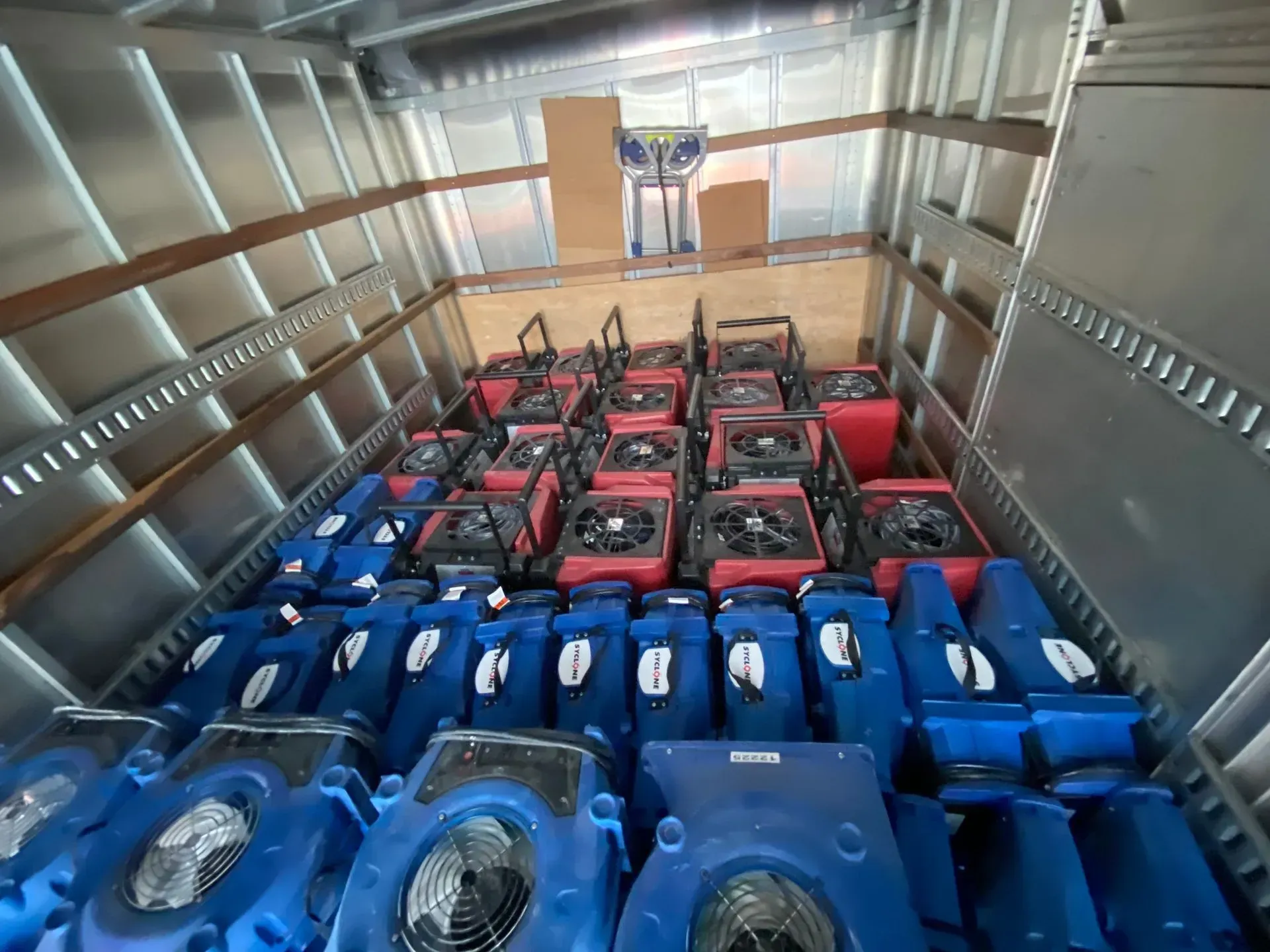 A box truck cargo area filled with rows of blue industrial air movers and several red industrial heaters.