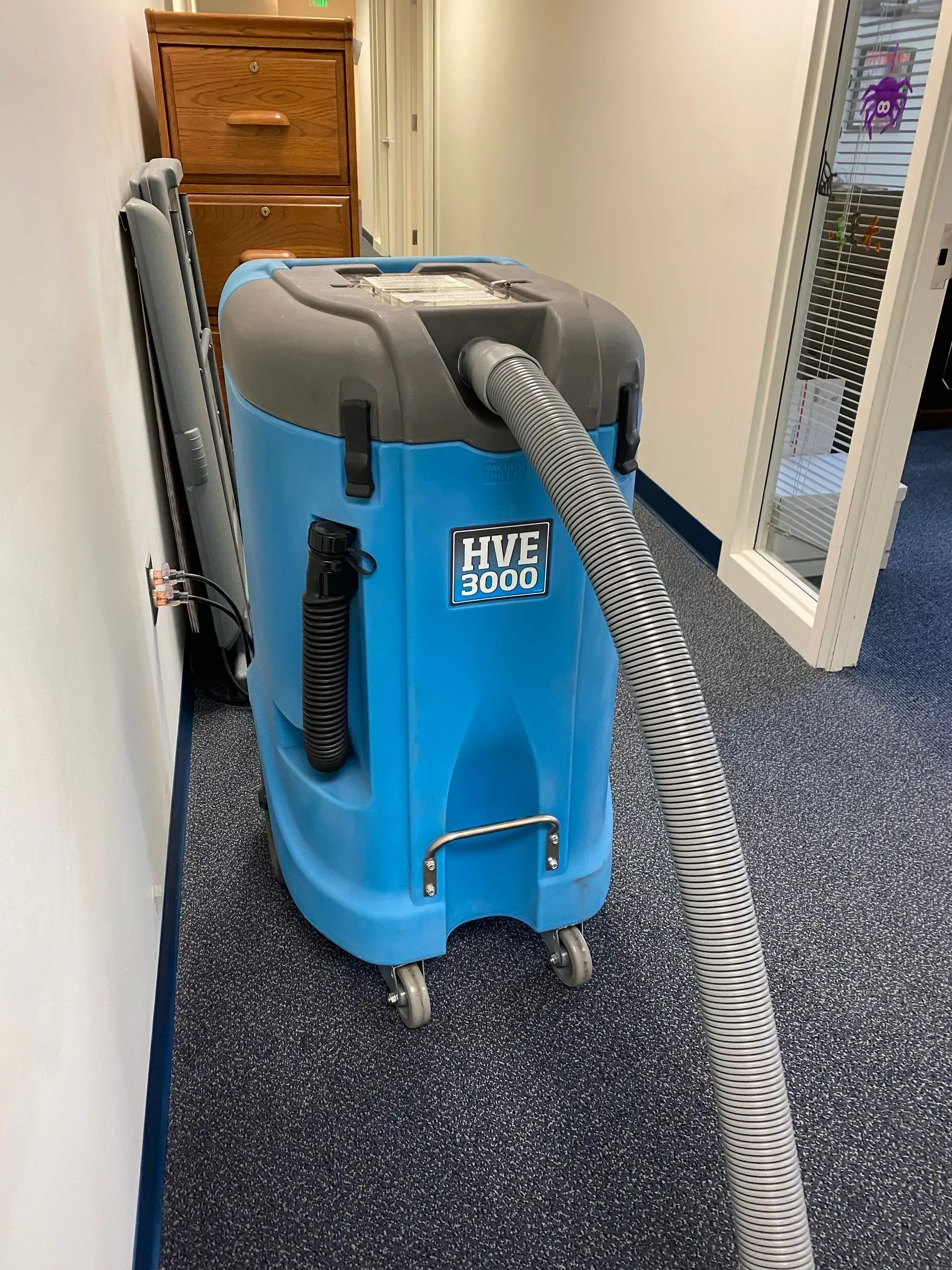 A blue HVE 3000 professional carpet cleaning machine with a grey hose stands on a speckled carpeted office floor.