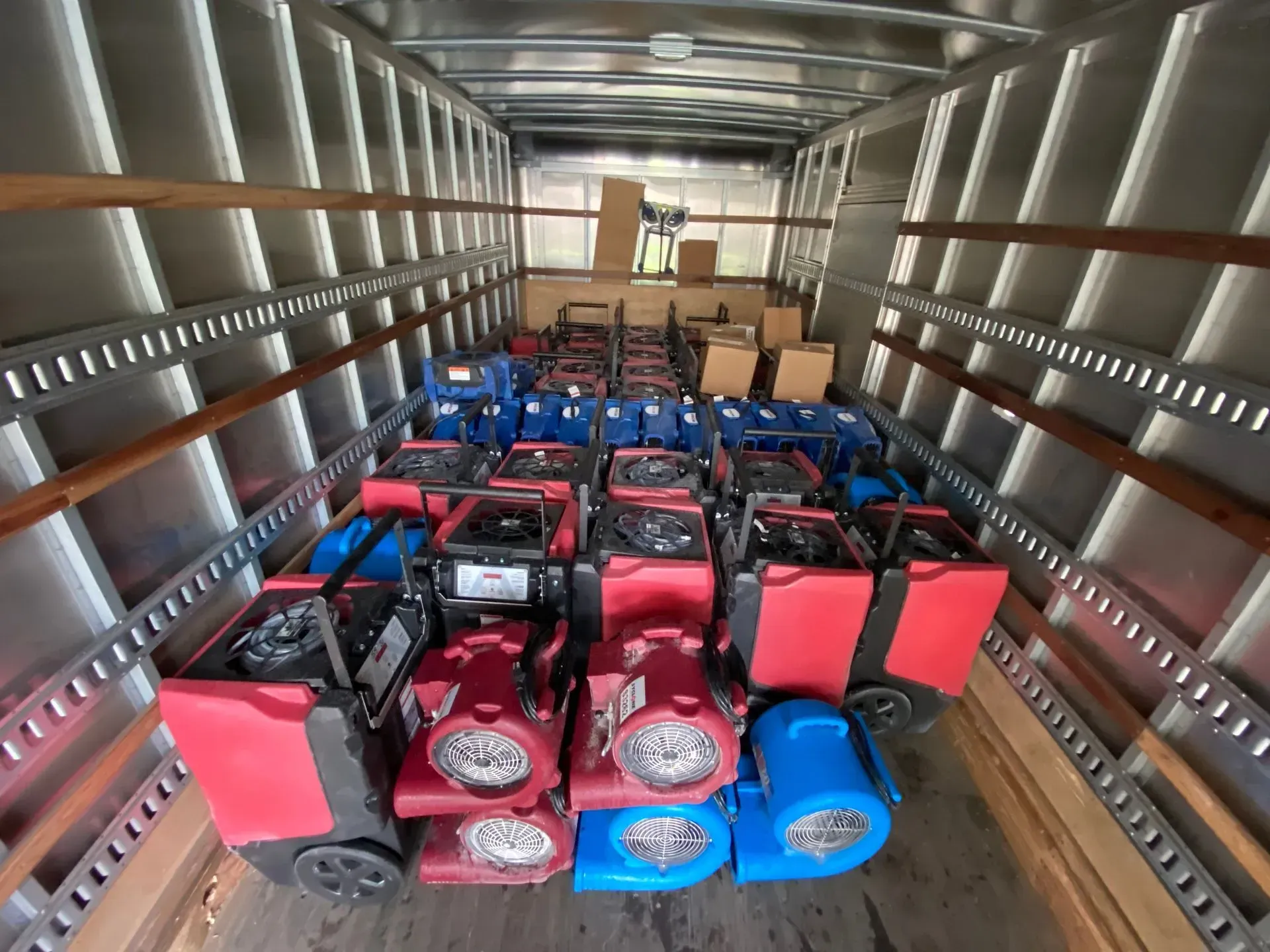 A cargo truck loaded with rows of red industrial dehumidifiers and blue air movers, with cardboard boxes in the back.