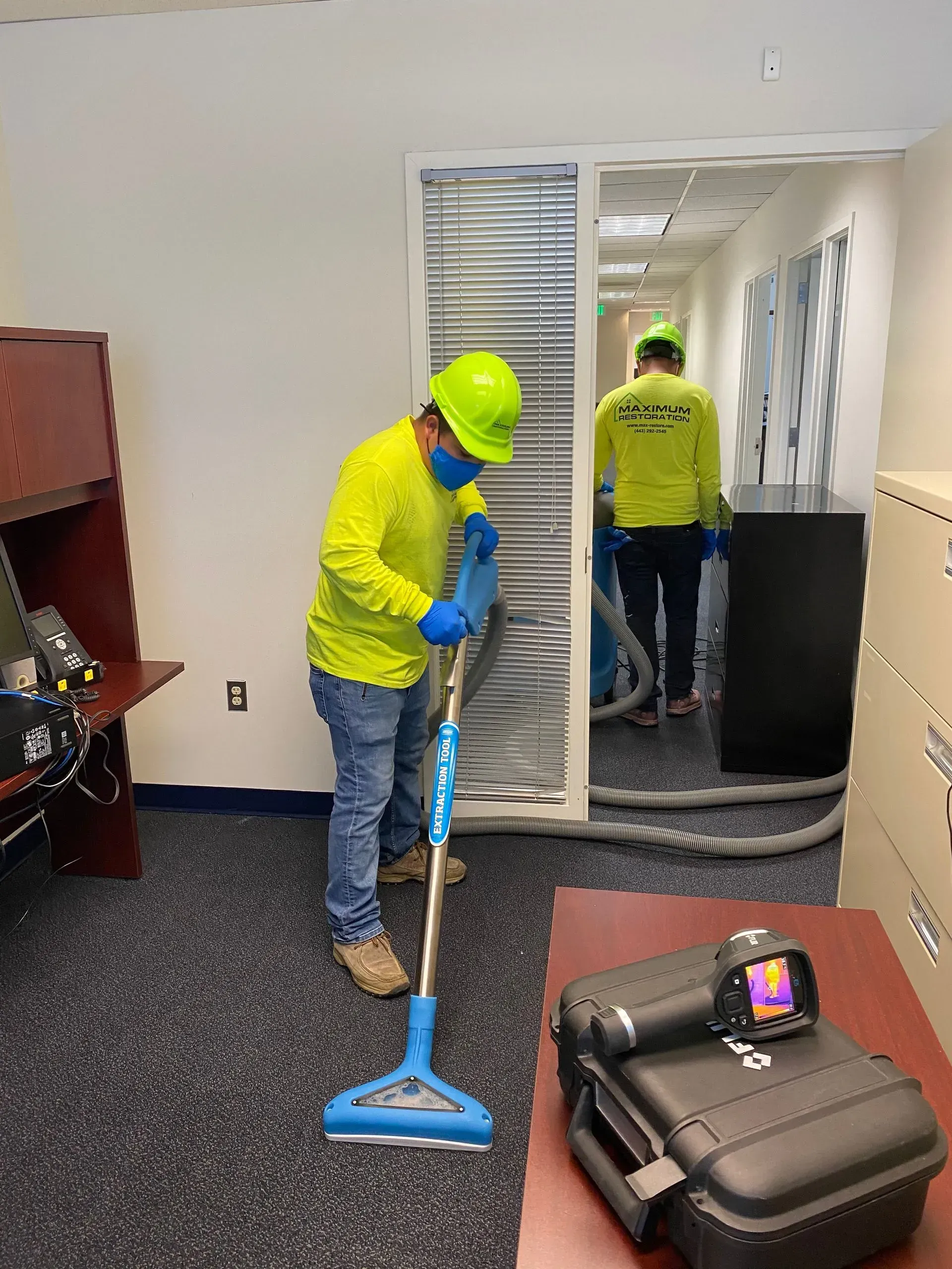Two workers in neon yellow shirts and hard hats clean a carpeted office using a specialized extraction vacuum wand.