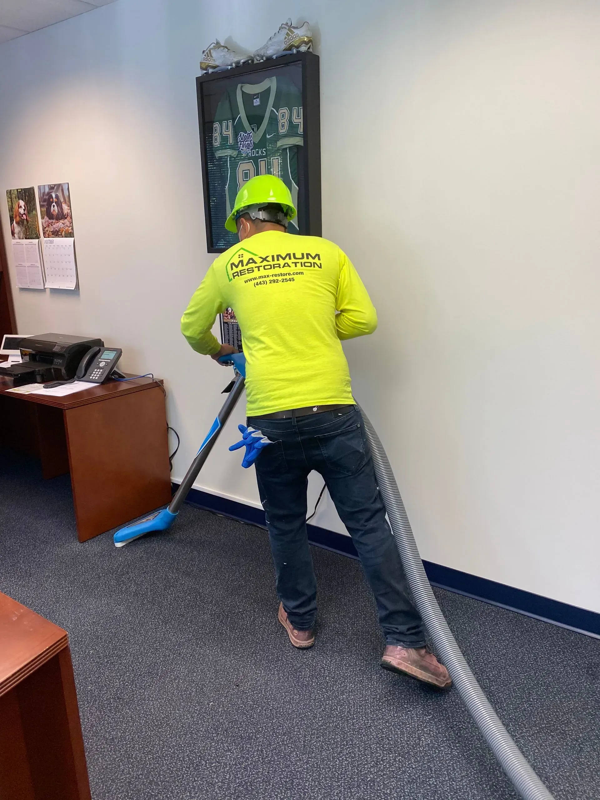 A worker in a neon yellow shirt and hard hat uses a carpet cleaning machine in an office setting.