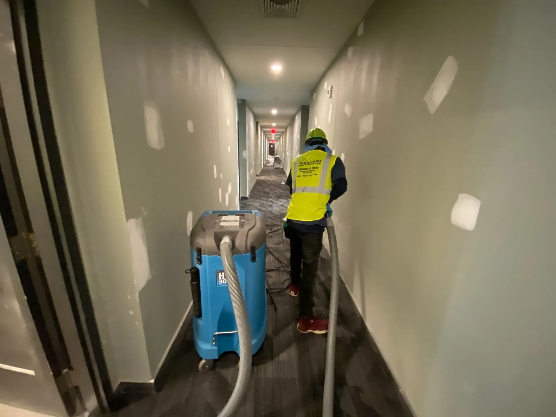 A worker in a high-visibility vest uses a blue industrial vacuum cleaner in a hallway with unfinished drywall patches.