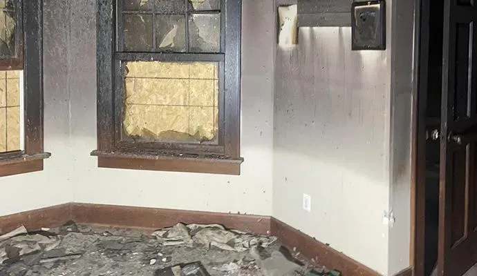 A room showing fire damage with soot-stained white walls, charred window frames, and debris scattered across the floor.