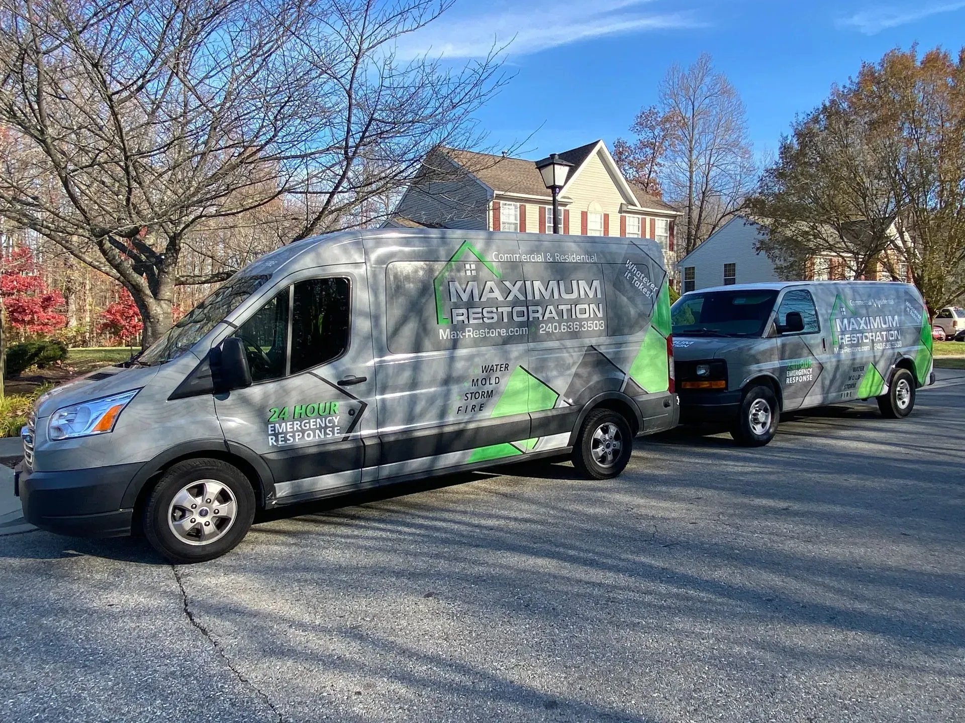 Two gray service vans parked on an asphalt driveway with green branding for Maximum Restoration on their sides.