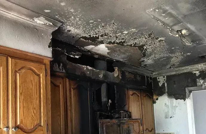Kitchen cabinets and ceiling charred from fire damage with peeling paint and heavy soot accumulation.