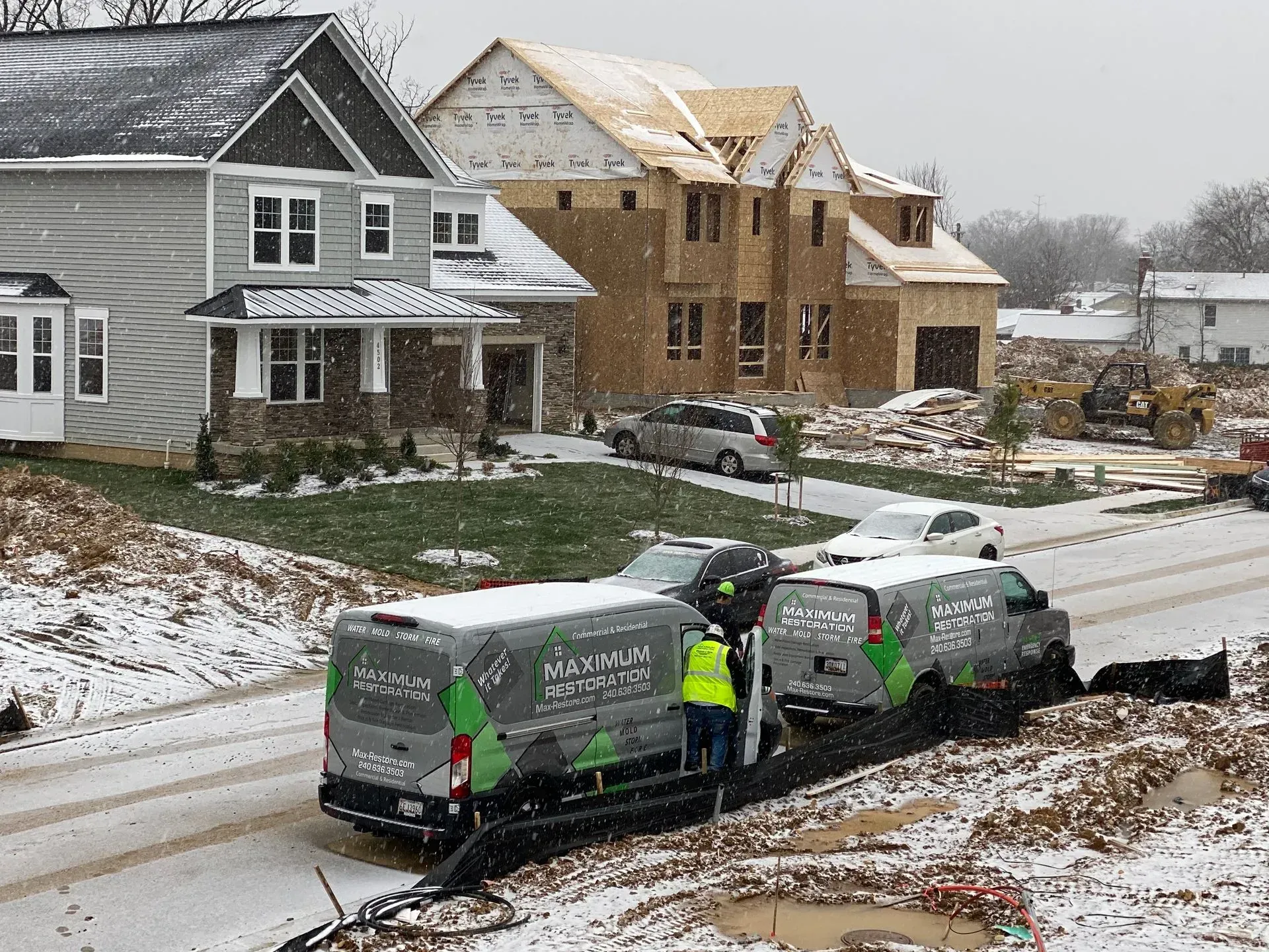 Construction workers in high-vis vests stand by grey utility vans on a snowy residential street near new homes.