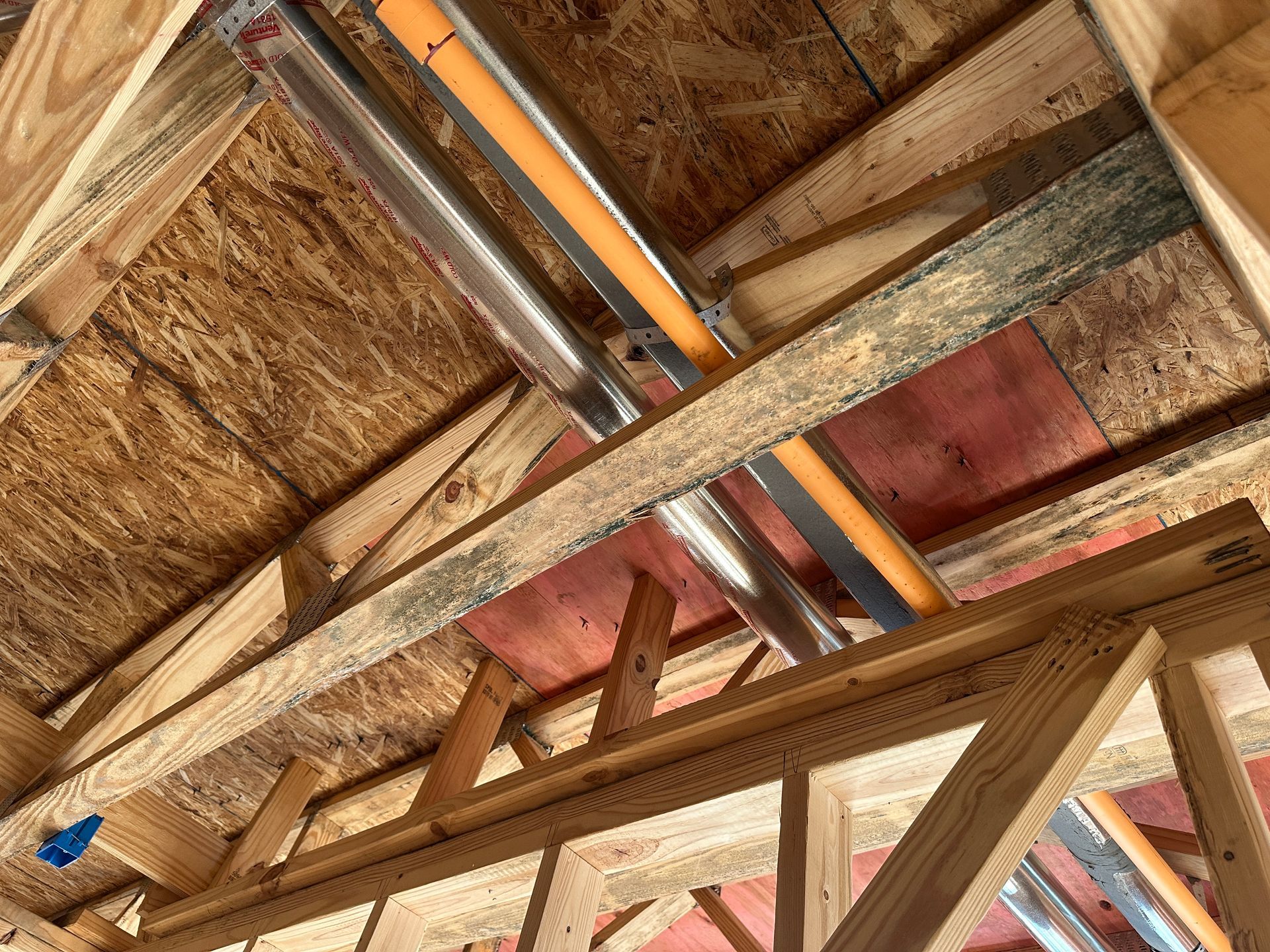 An attic view showing wooden roof trusses and structural beams with metal and yellow pipes running along the rafters.