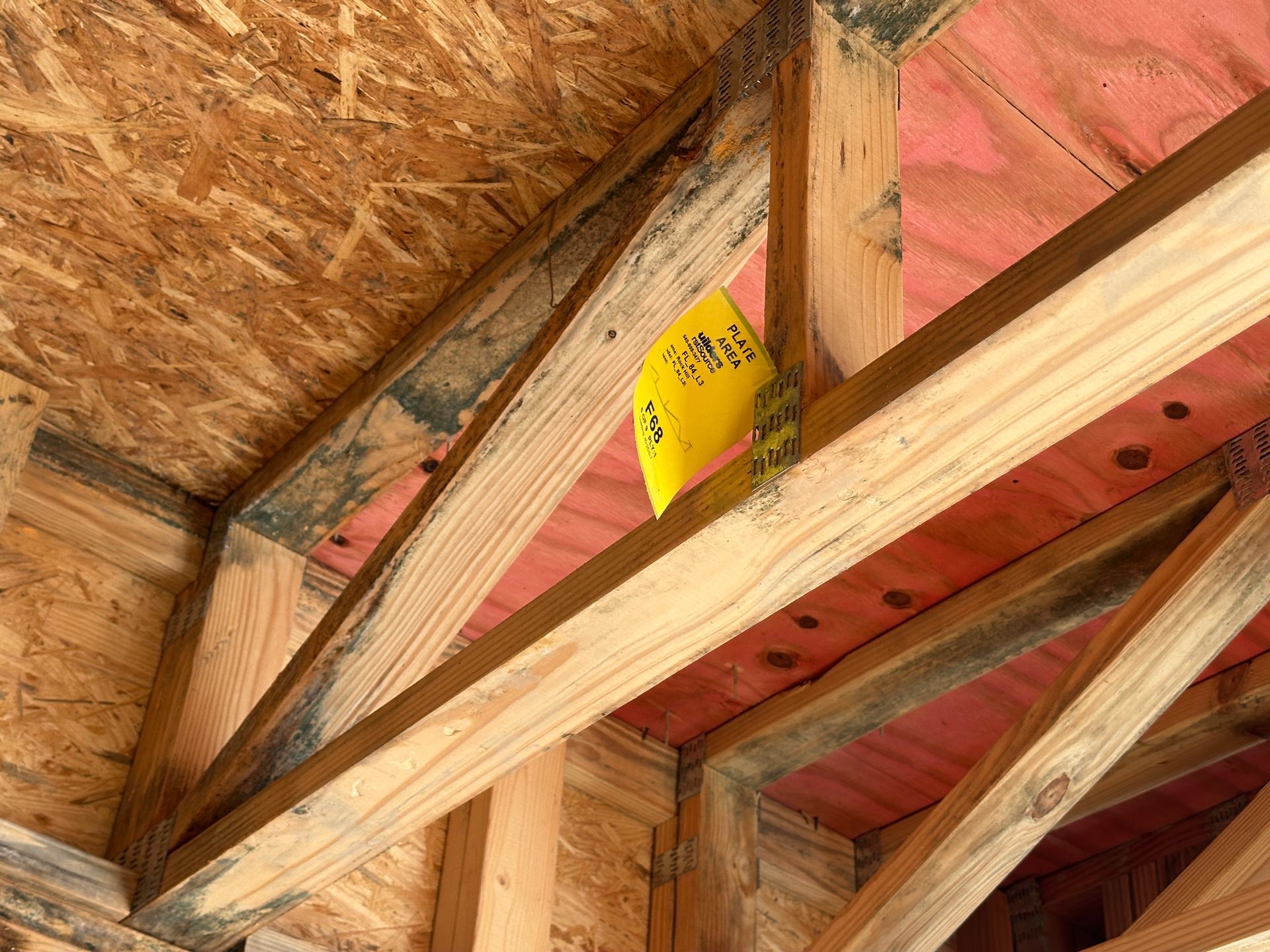 A yellow inspection tag clipped to wooden roof trusses and OSB sheathing in an unfinished attic space.