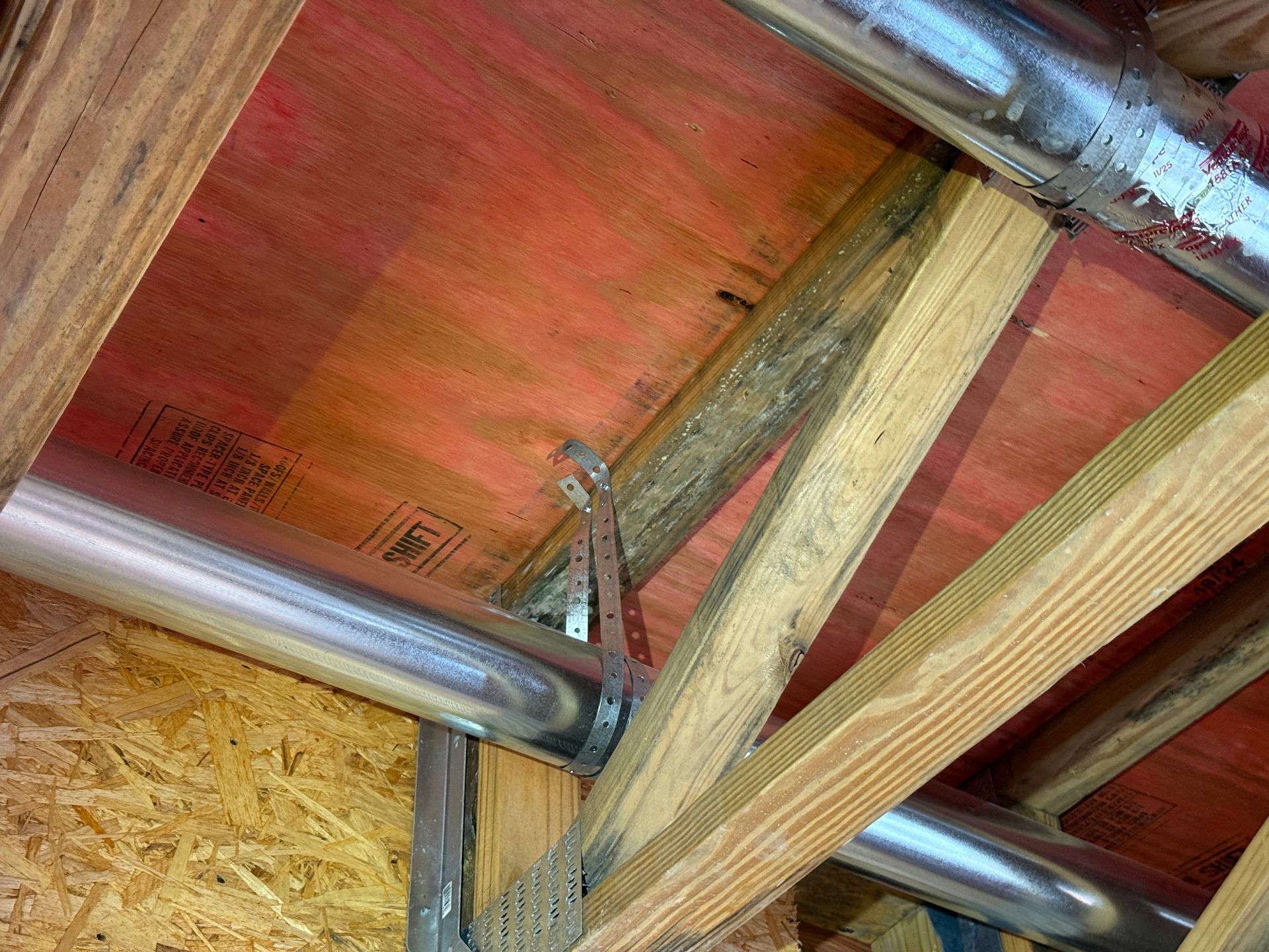 Metal ductwork suspended by metal straps from wooden ceiling rafters in an unfinished attic space.