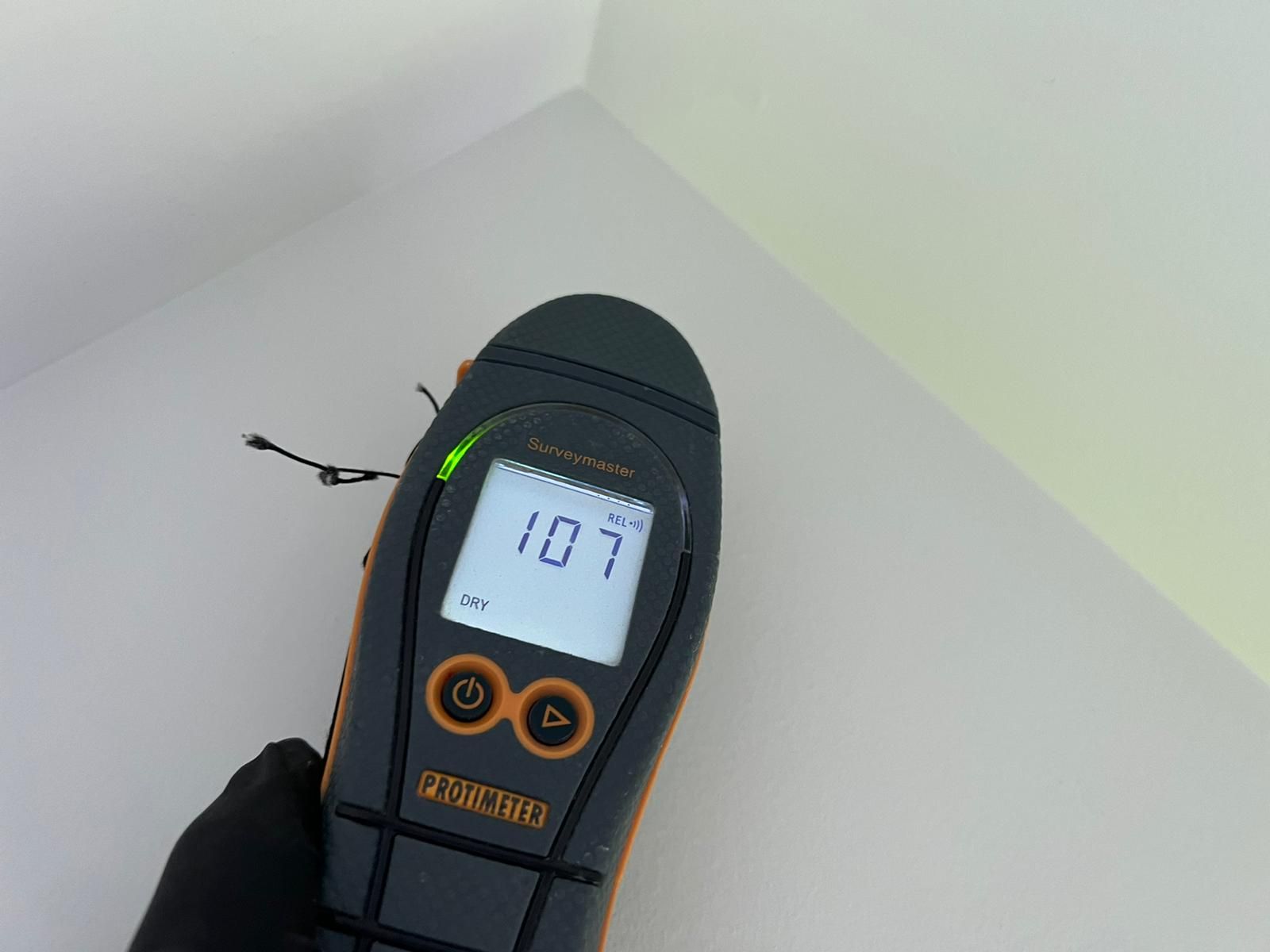 A person using a moisture meter to check a white wall in a corner. The meter reads 107.
