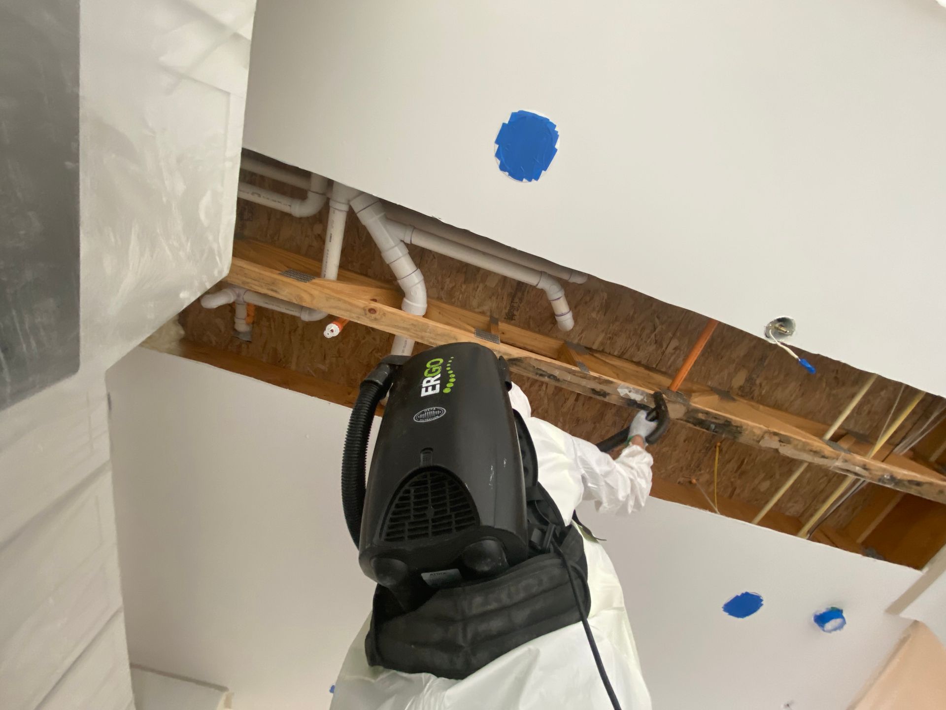 Person in protective suit removing ceiling material, backpack vacuum. Exposed plumbing, blue paint marks.