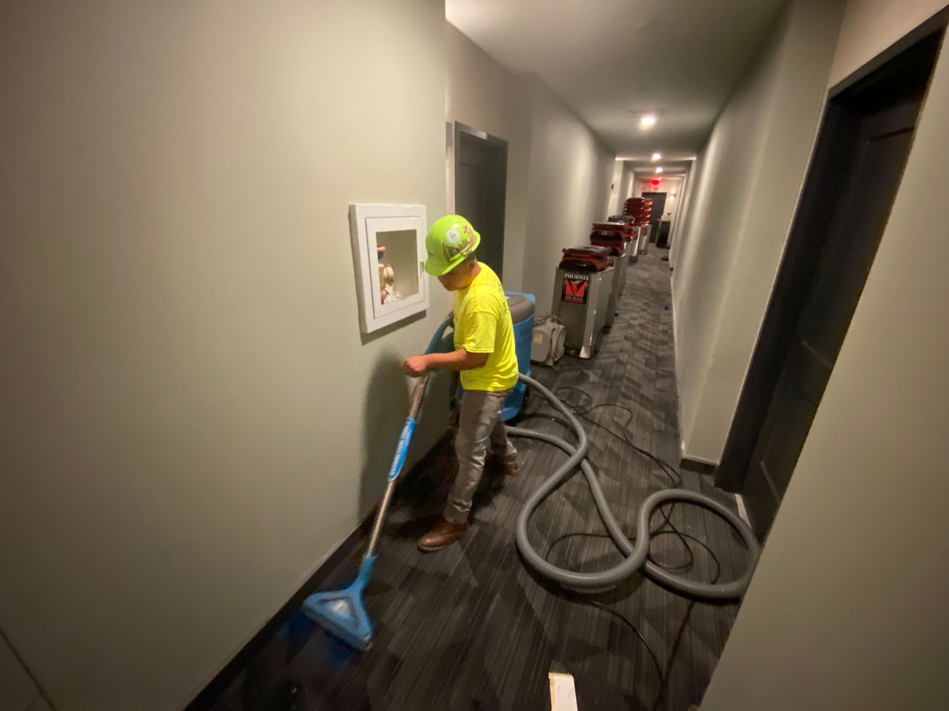 Person in a yellow shirt and hard hat vacuuming a hallway carpet with several machines.