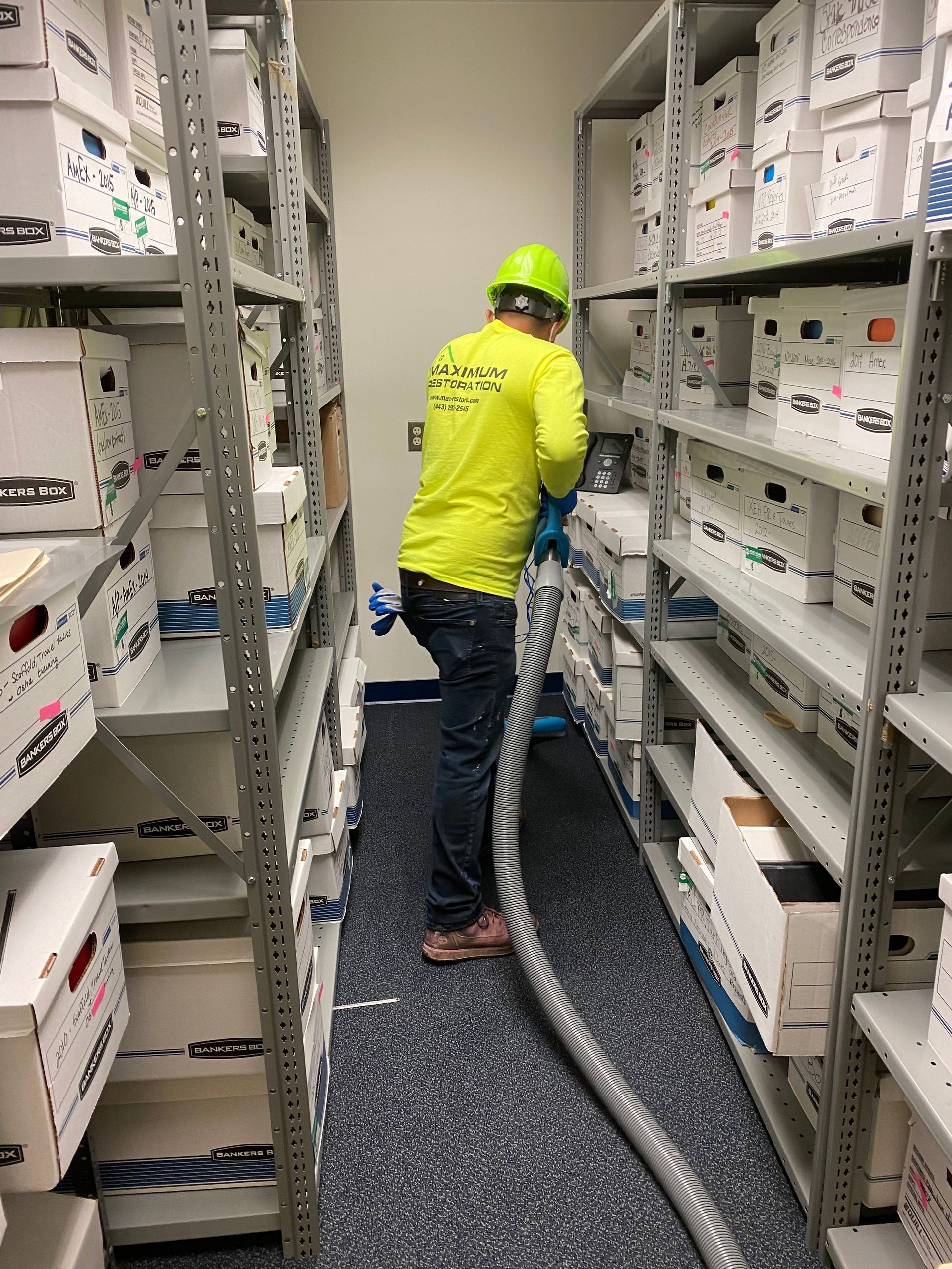Person in a yellow shirt and green hardhat vacuums between shelves filled with boxes in a storage room.