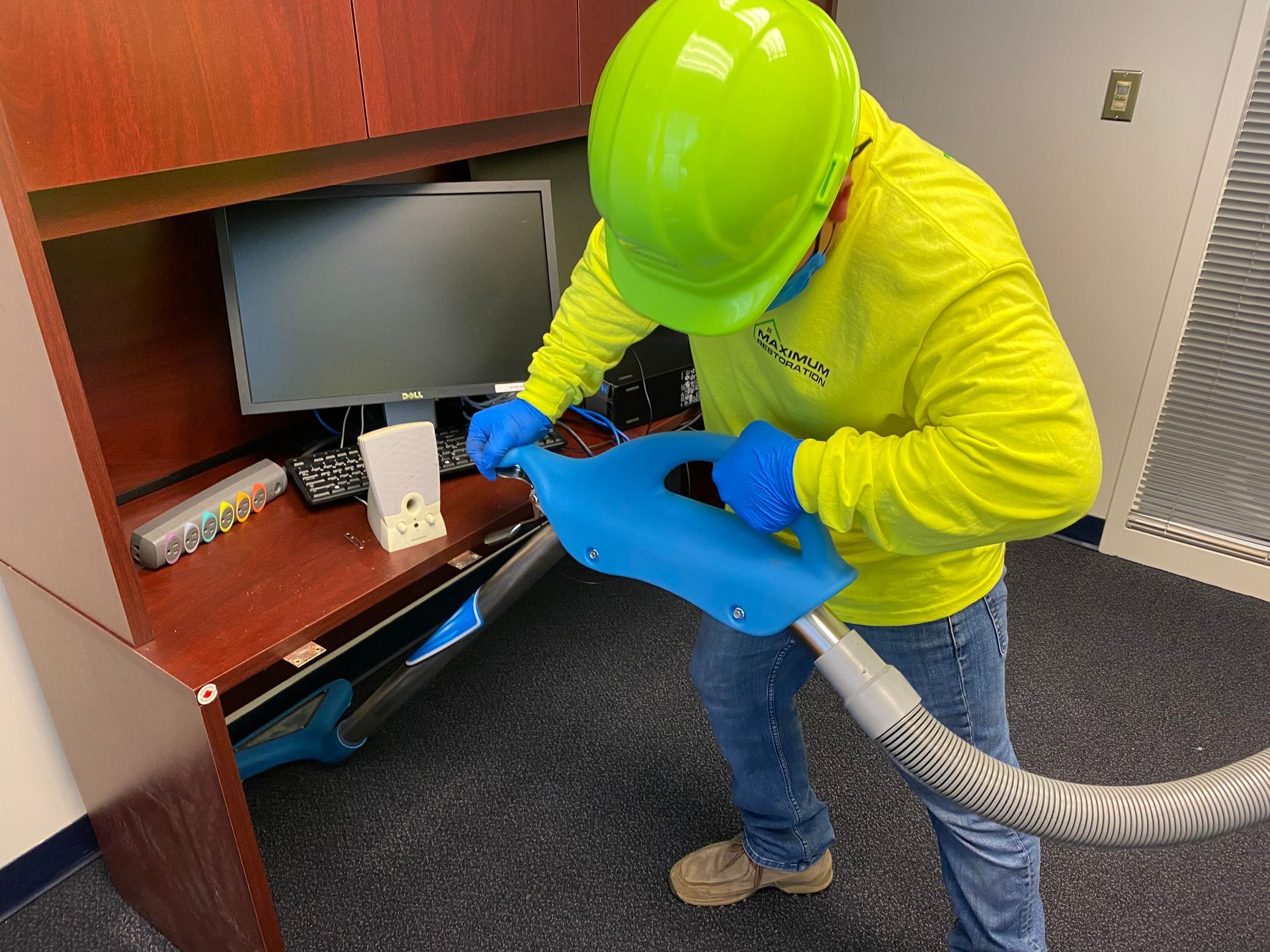 Person in hard hat and gloves cleaning an office desk with a vacuum hose.