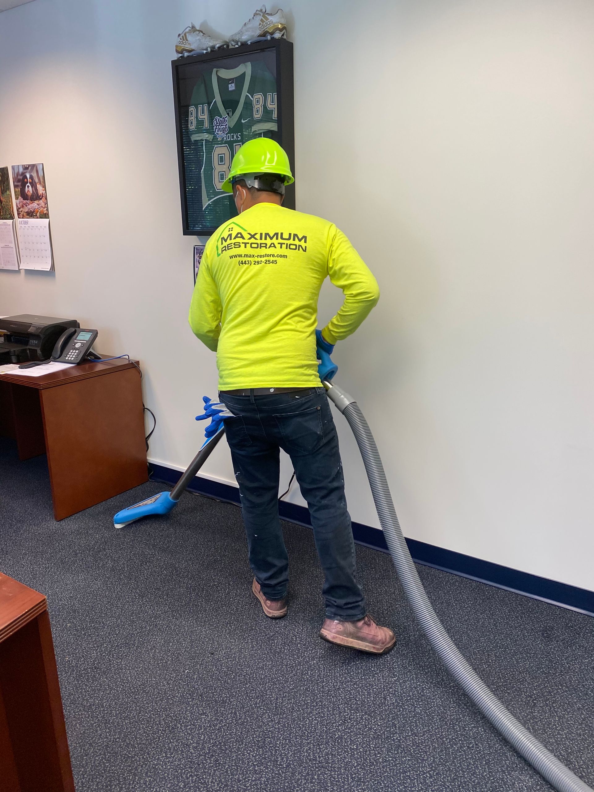 Person in yellow shirt vacuums office carpet, wearing a hard hat.