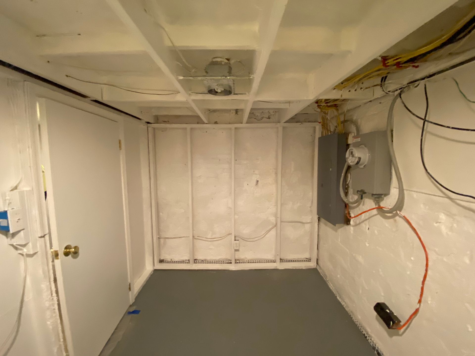 Basement room with white walls, gray floor, electrical panel, door, and exposed ceiling beams.