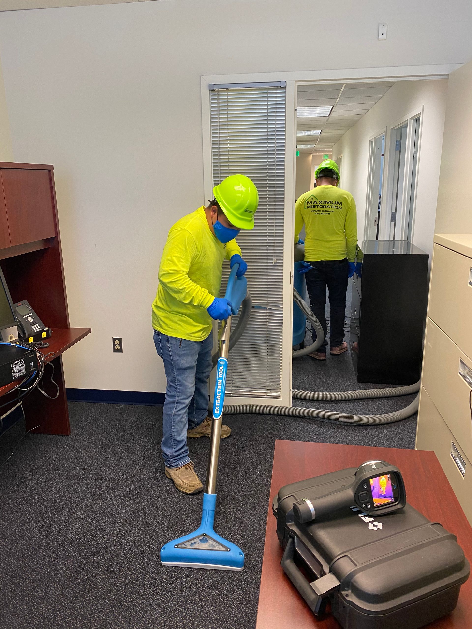 Two people in safety gear vacuuming carpet in an office. One vacuums near a desk; the other walks down the hallway.