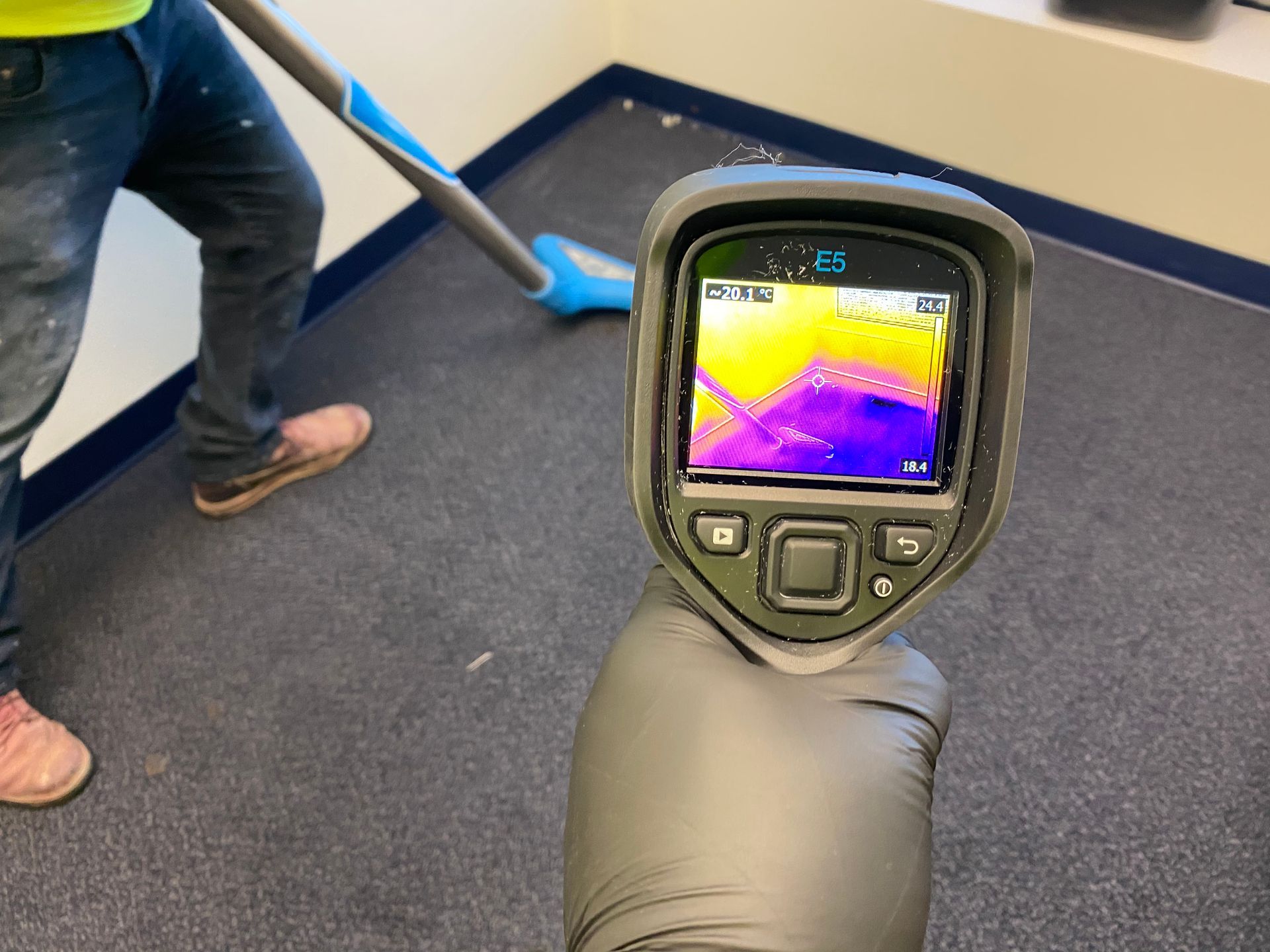 Hand holding thermal camera pointed at a carpeted floor. A person vacuums in the background. Camera display shows purple and yellow heat signatures.