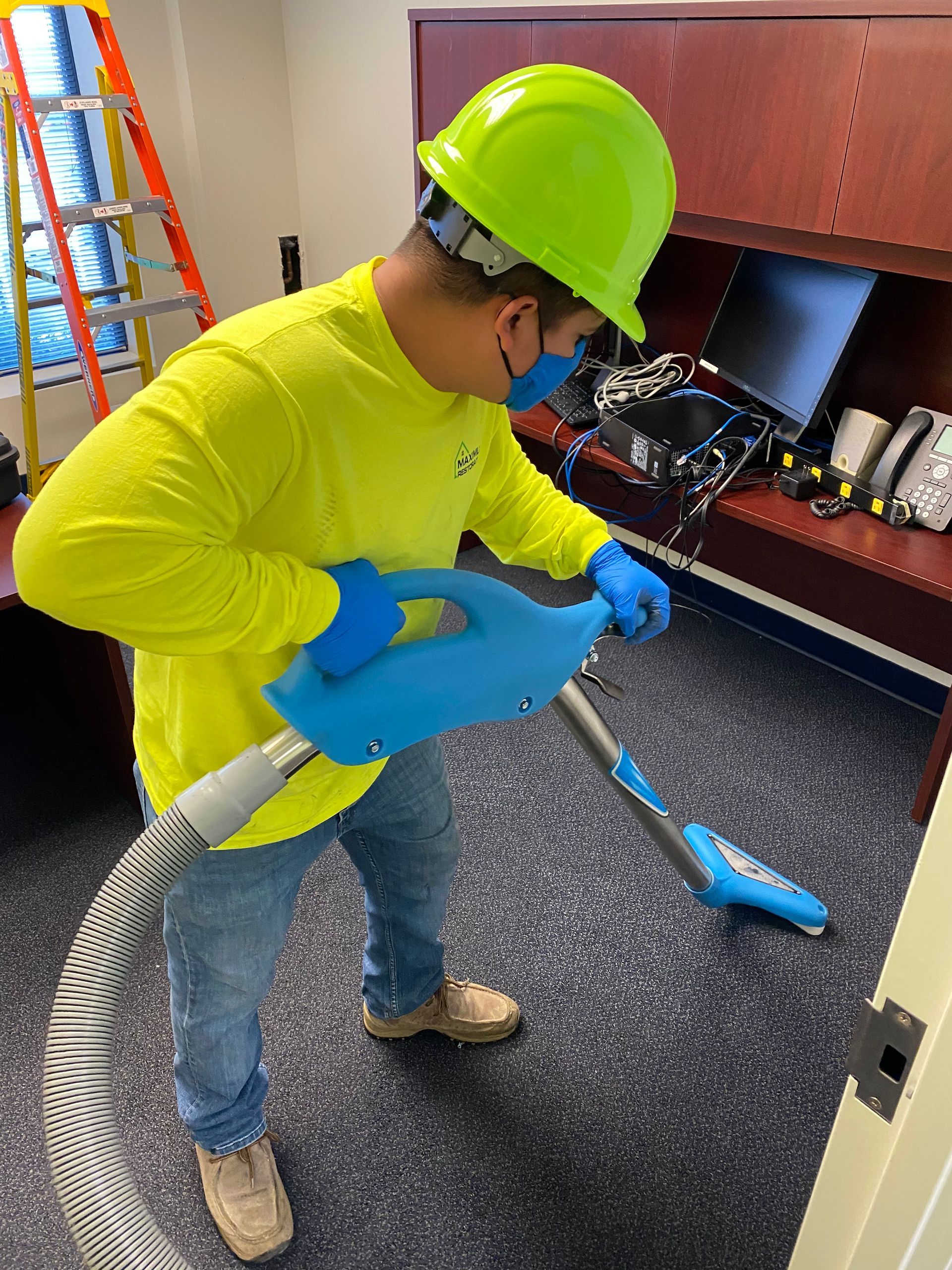 Person in hard hat, gloves, and mask vacuuming a carpeted room.