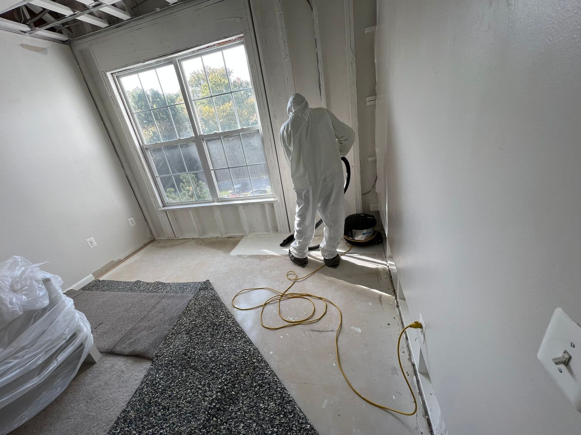 Person in protective suit cleaning a room with a window; debris and vacuum present.