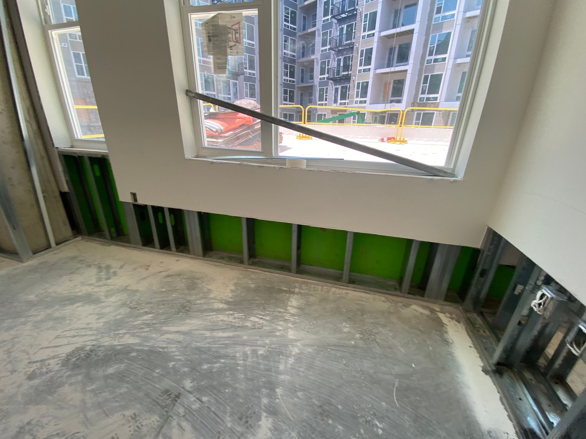 Interior construction site with metal studs, green insulation, and a window overlooking another building.
