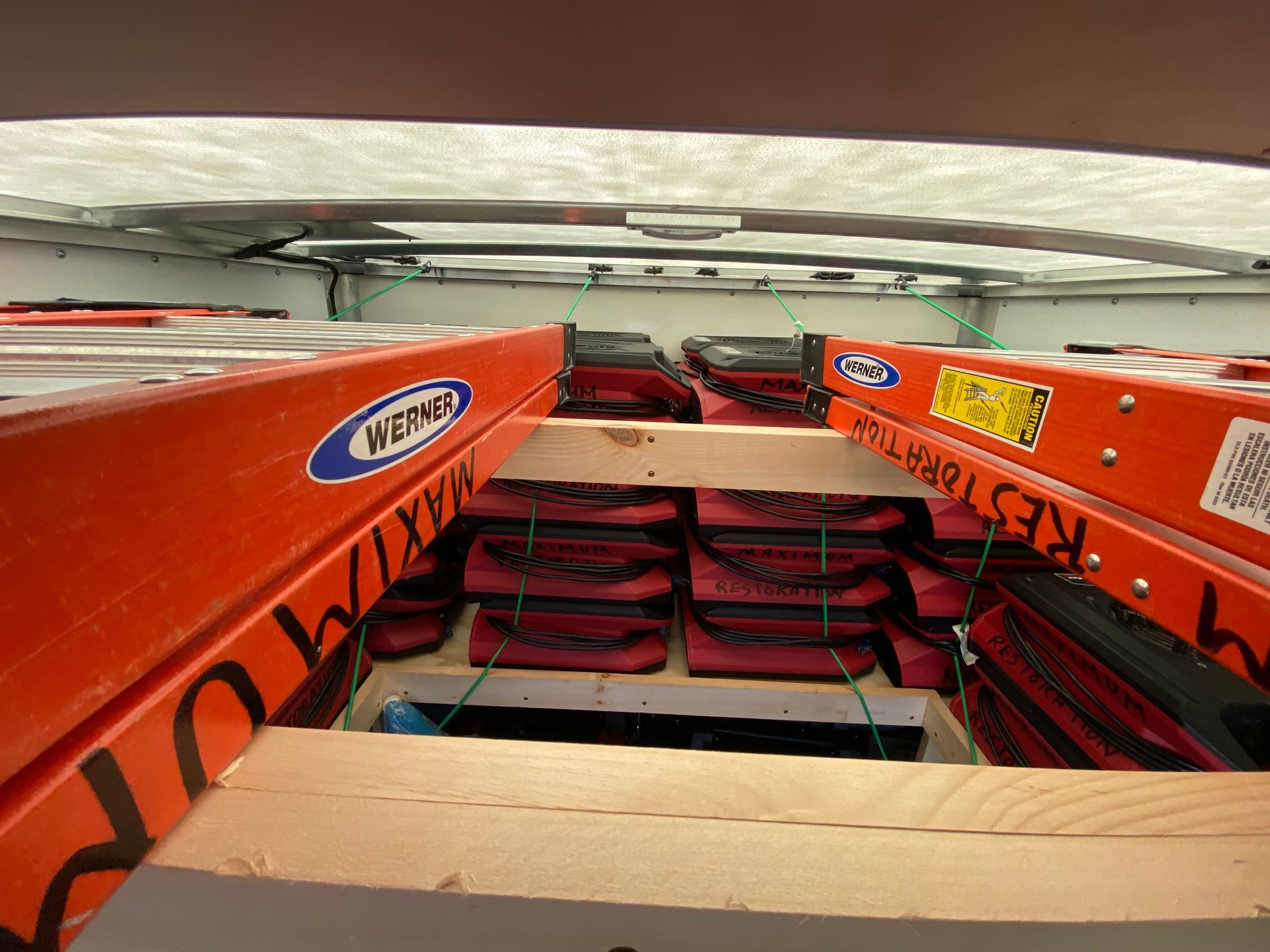 Inside a van: two orange ladders atop red bags, secured with green cords. Wooden supports and a white roof.