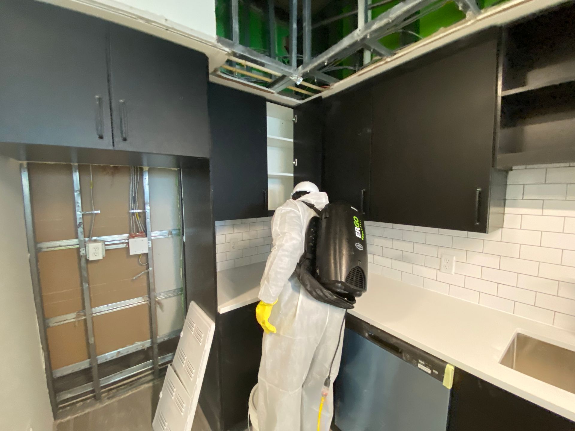 Person in protective suit disinfecting kitchen with black cabinets and white tile backsplash.