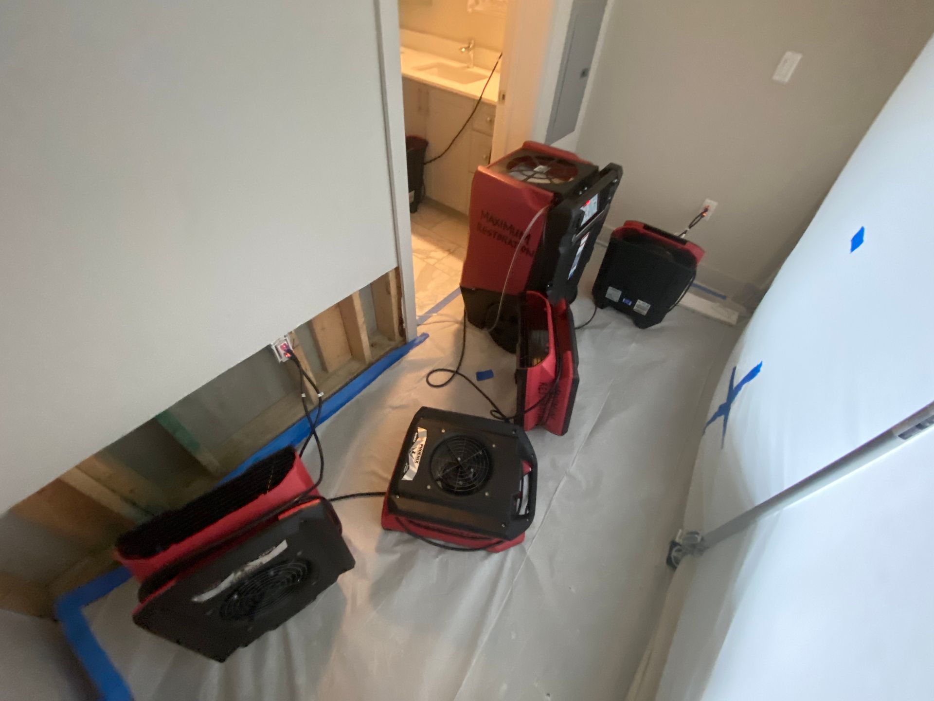 Water damage restoration equipment in a room with a section of drywall removed, tented with plastic sheeting.