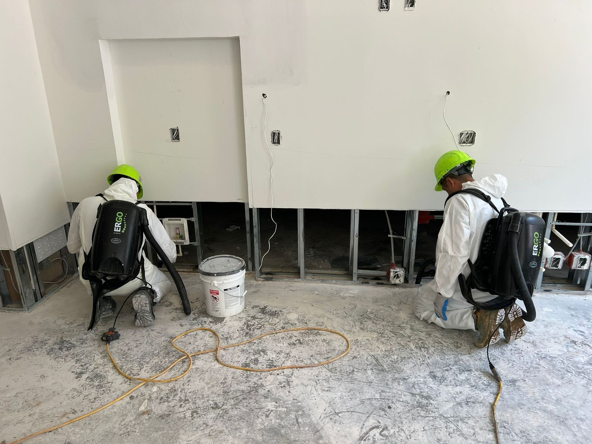 Two workers in protective suits inspect a wall, possibly for mold remediation, indoors.